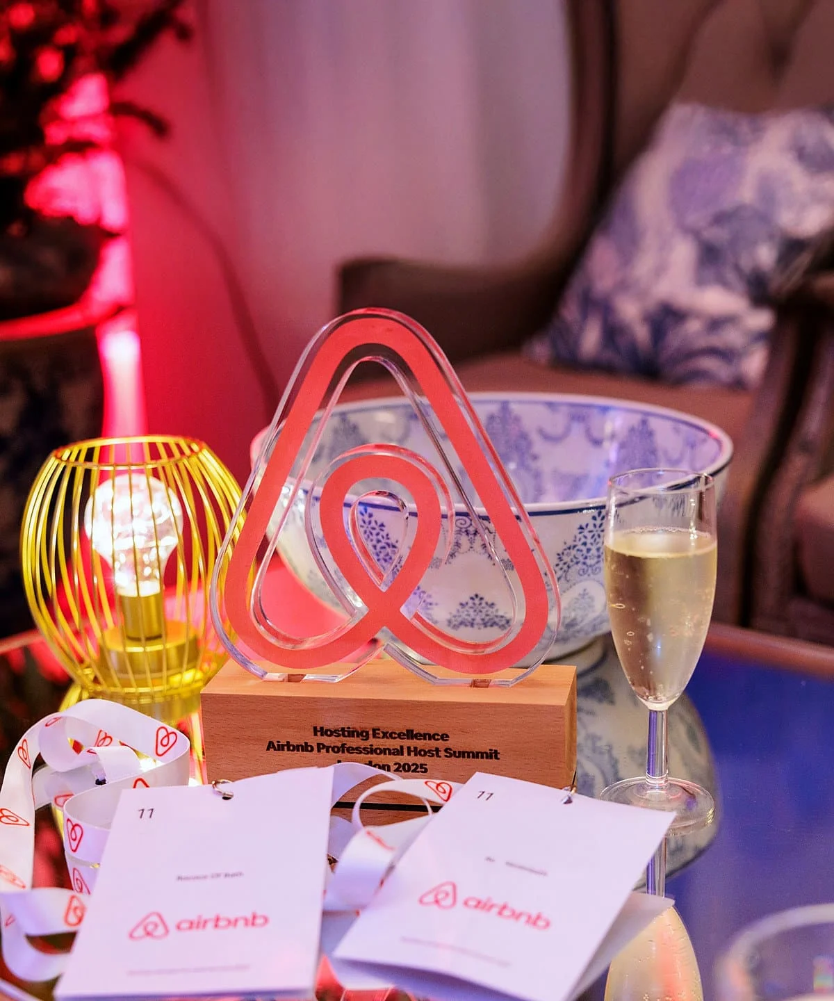 The Airbnb Hosting Excellence award from the Professional Host Summit London 2025, on a table at the event with an Airbnb lanyard and a glass of champagne.